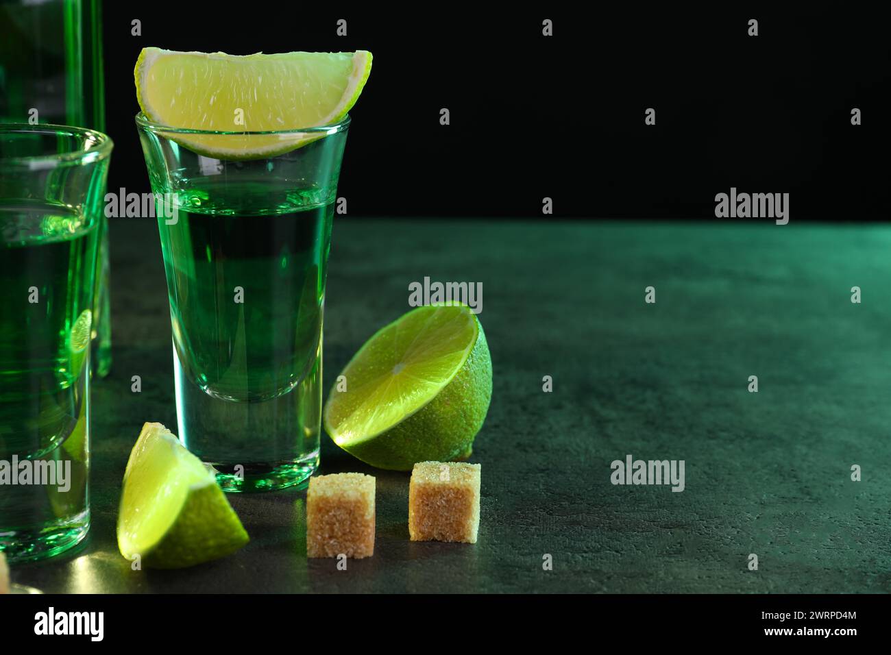 Absinthe in bicchieri da shot, cubetti di lime e zucchero di canna su un tavolo testurizzato grigio su sfondo nero, spazio per il testo. Bevanda alcolica Foto Stock