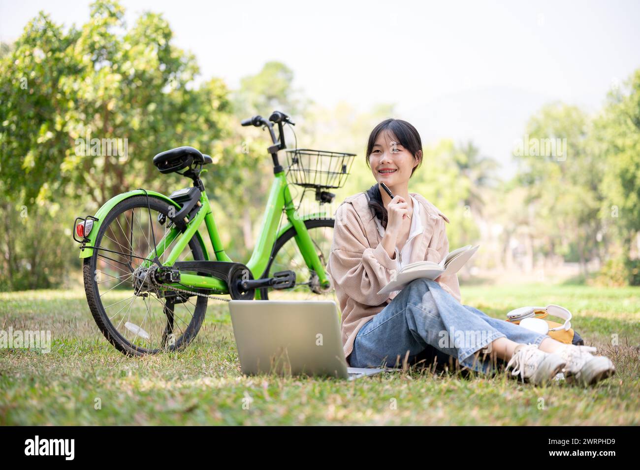 Una giovane studentessa asiatica, felice e positiva, sta facendo i compiti in un parco universitario, seduta sull'erba con il suo laptop, lo zaino e la bicicletta. Foto Stock