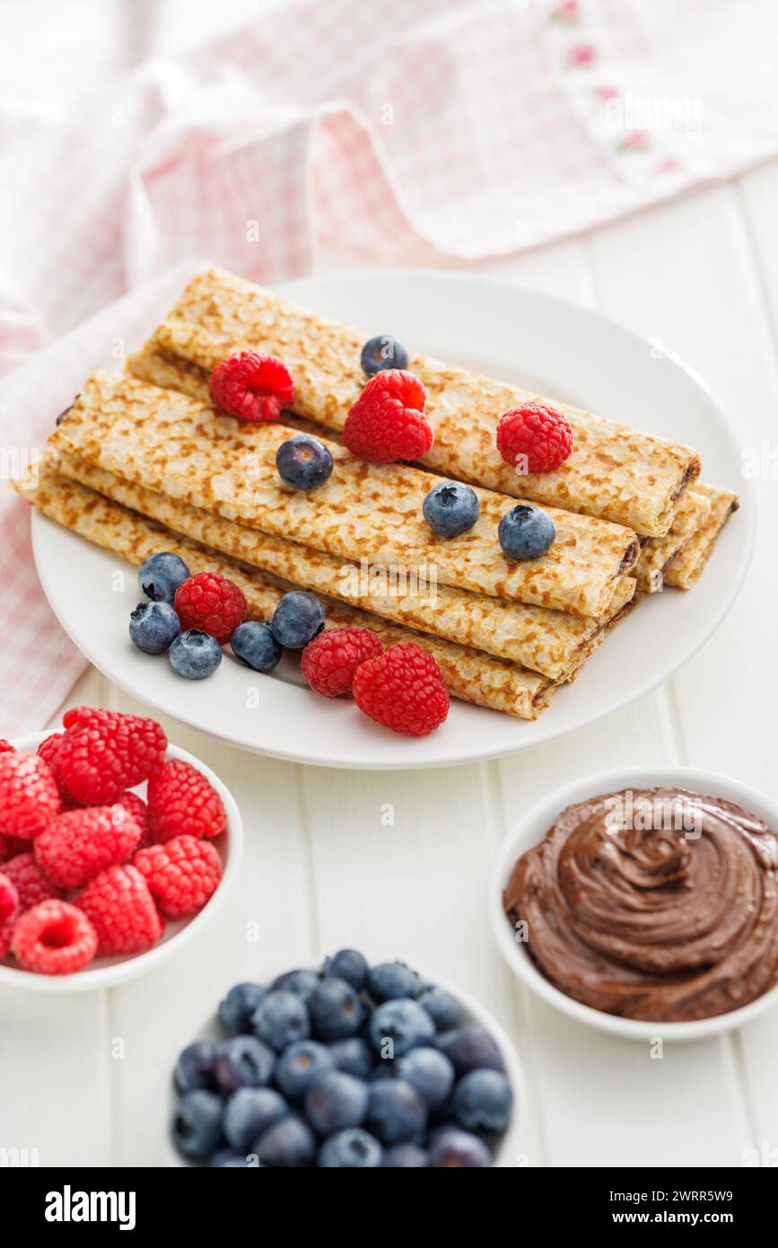 Crepe fresche ricoperte di frutti di bosco freschi servite su un piatto bianco accanto a ciotole di lamponi, mirtilli e spalmazione al cioccolato. Foto Stock