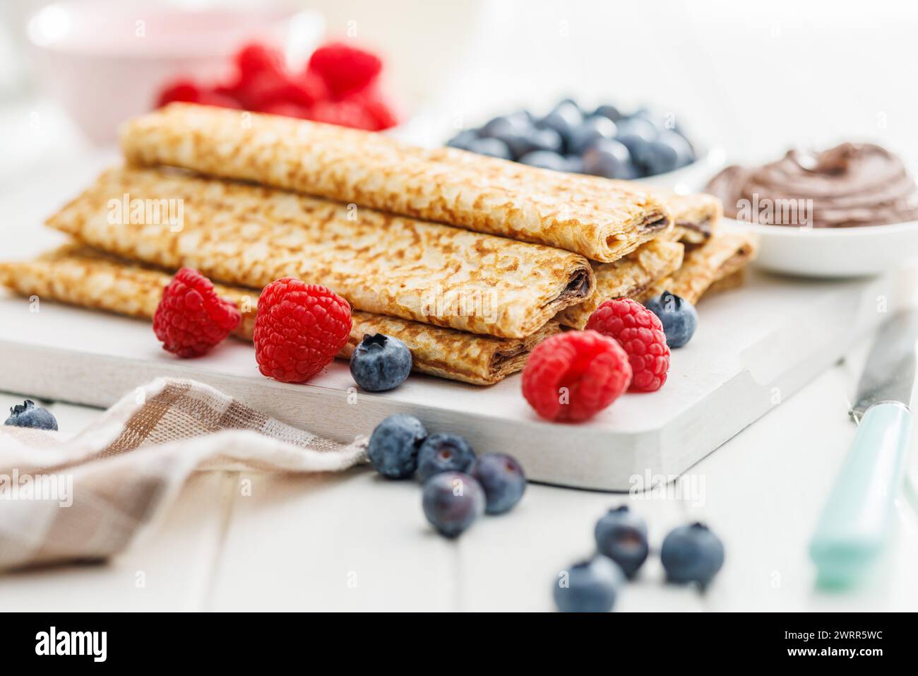 Crepe fresche ricoperte di frutti di bosco freschi servite su un piatto bianco accanto a ciotole di lamponi, mirtilli e spalmazione al cioccolato. Foto Stock