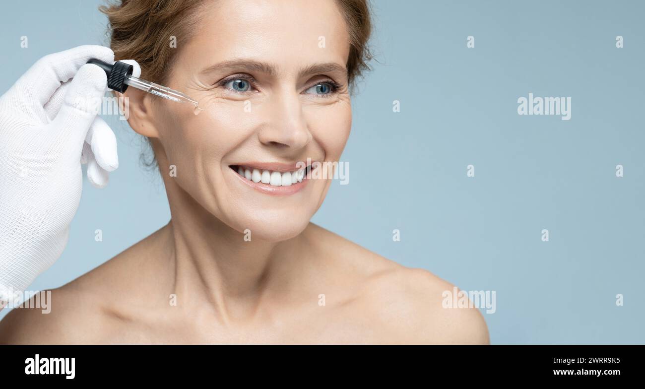 Il cosmetologo che utilizza il contagocce per pipette applica l'olio essenziale al viso di una donna matura per una pelle idratata. Cura della pelle del viso con olio essenziale per il trattamento e. Foto Stock