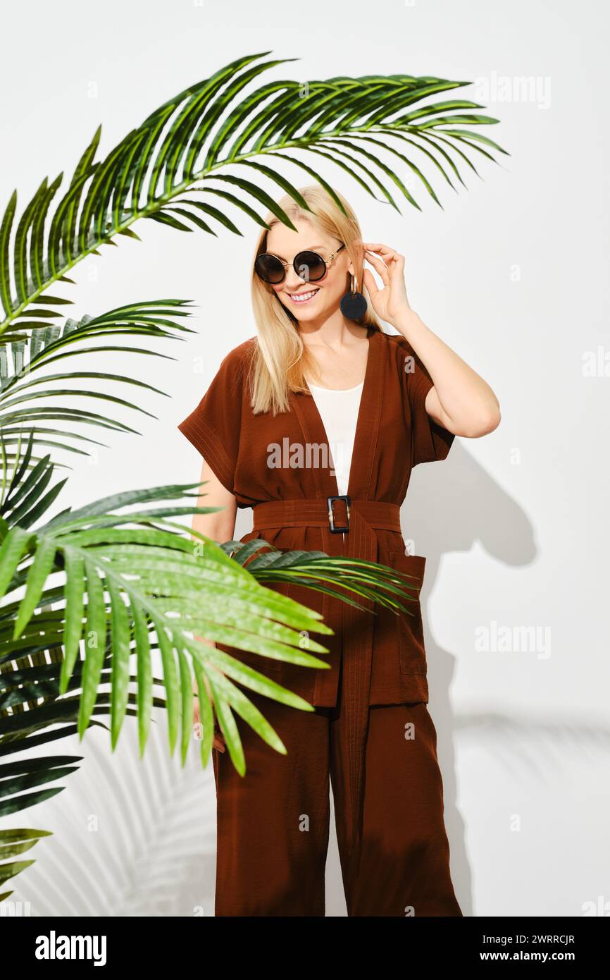 Donna felice in tuta marrone che si posa dietro a foglie di palma verde vibrante creando un contrasto dinamico ed esteticamente piacevole Foto Stock