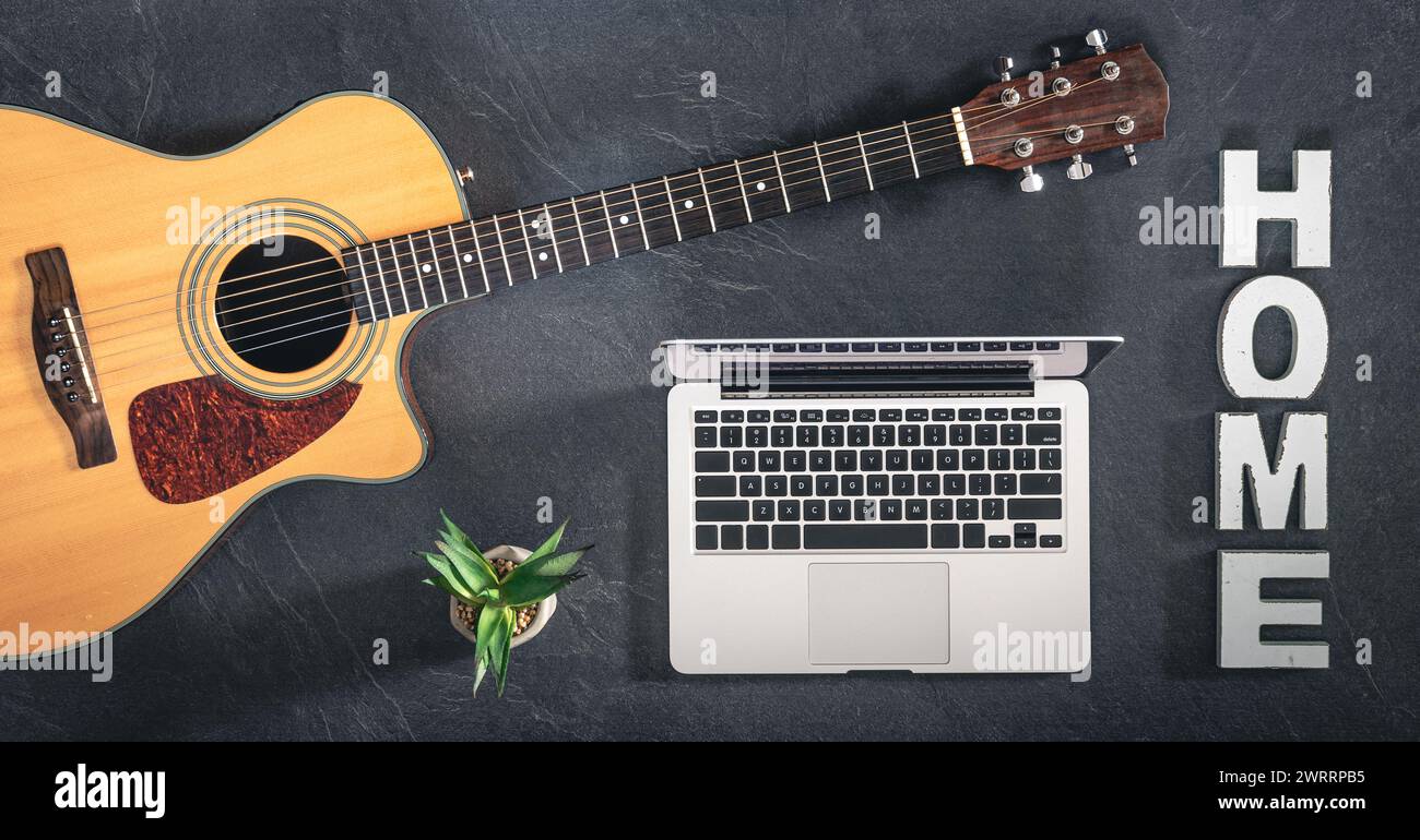Chitarra acustica, laptop e parola decorativa Home su sfondo scuro, vista dall'alto. Foto Stock