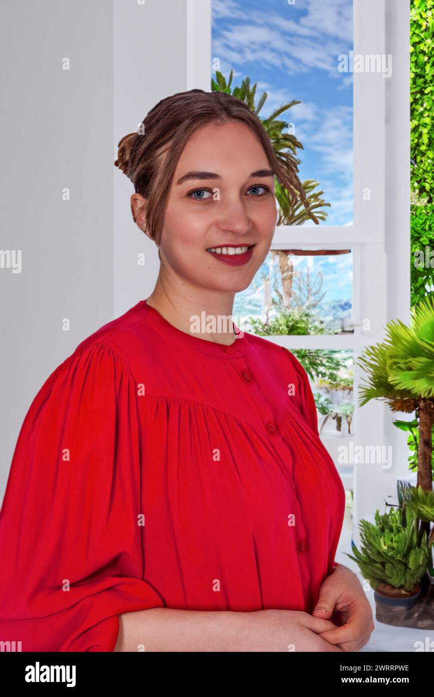 La foto mostra la parte superiore del corpo di una giovane donna in una camicetta rossa brillante seduta di fronte a una finestra, sorridendo allegramente. La luce naturale lo mette in risalto Foto Stock
