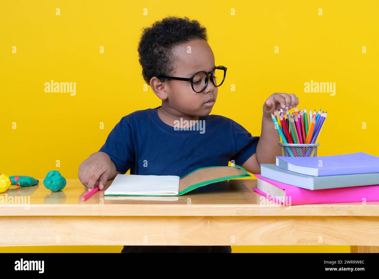 Bambino con immaginazione seduto al tavolo disegnando con matite colorate su sfondo giallo. Foto Stock