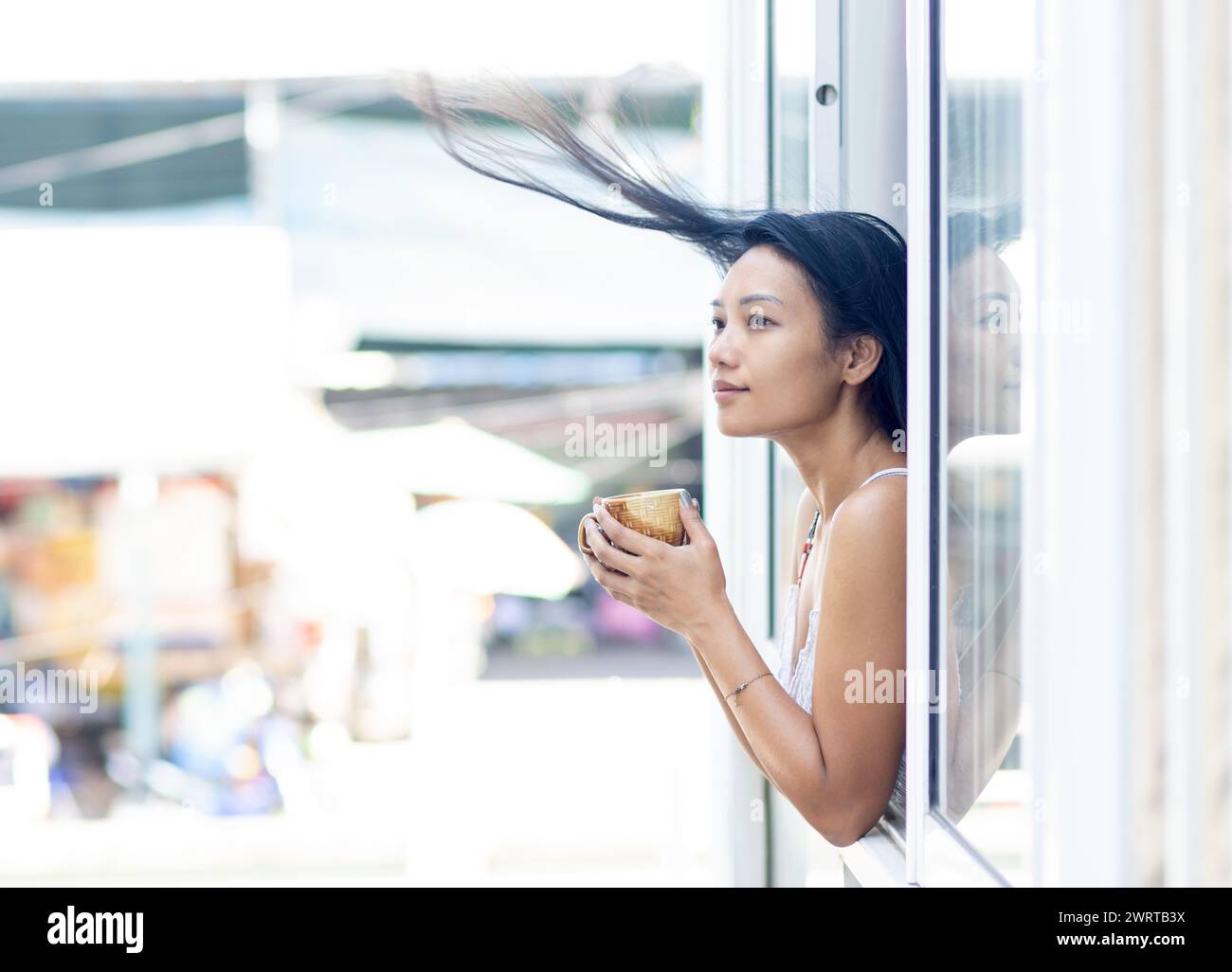 Una donna felice in un abito estivo tiene una tazza in una finestra aperta di un edificio residenziale mentre il vento le soffia i capelli Foto Stock