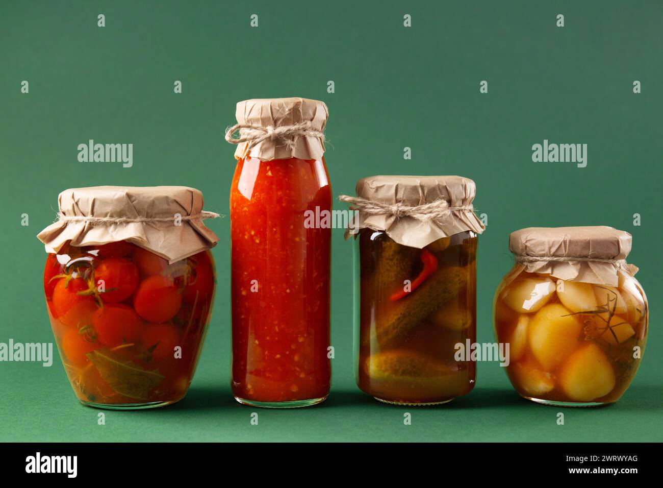 Verdure in scatola in vasetti di vetro chiusi su sfondo verde. Cetrioli sottaceto, pomodori, salsa al peperoncino, aglio, adjika. Spuntino piccante, cibo in scatola. Foto Stock