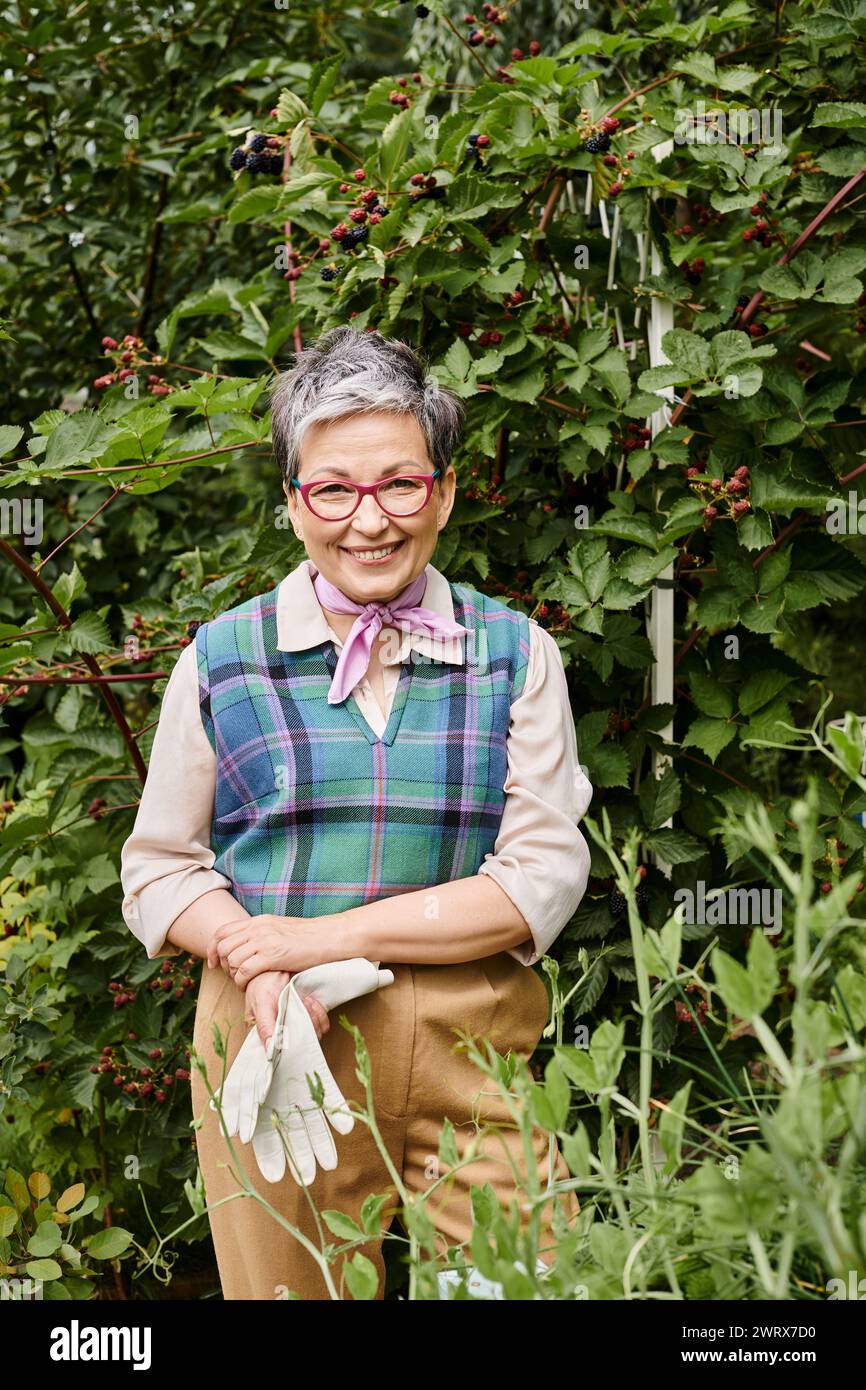 donna matura, gioiosa e raffinata, con guanti e occhiali che si prende cura delle sue piante e sorride alla macchina fotografica Foto Stock