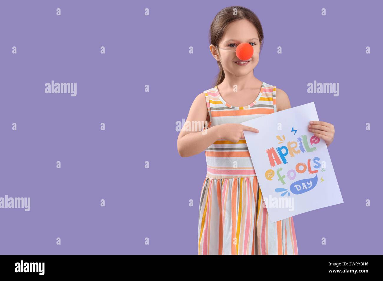 Una bambina carina con il naso da clown che punta al poster con il testo APRIL FOOLS DAY su sfondo lilla. Festa del giorno dello scemo d'aprile Foto Stock