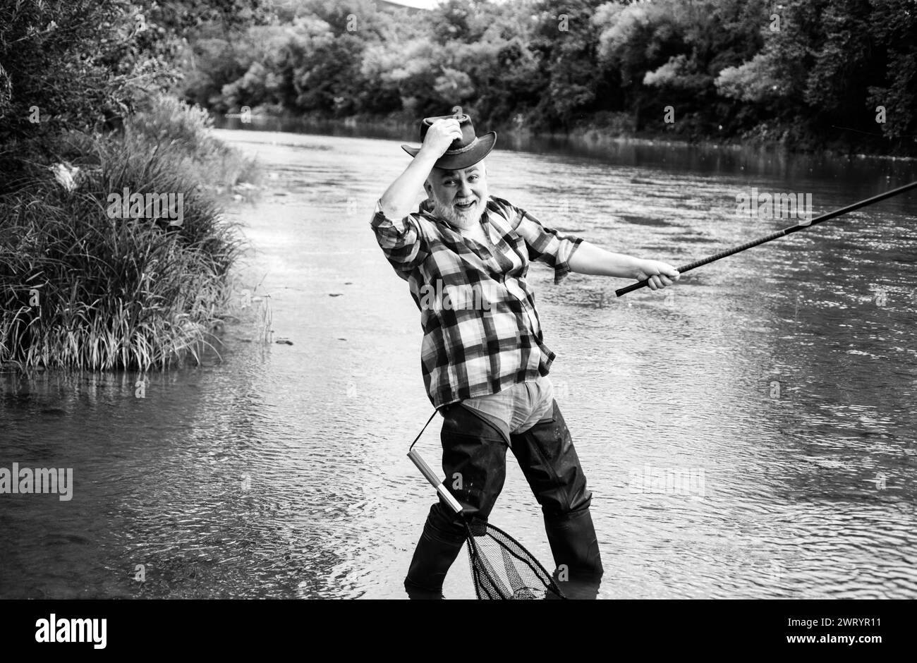 Pescatore esperto eccitato pescatore in cappello cowboy con canna da pesca, bobina rotante sul fiume. Vecchio uomo che cattura il pesce, tirando la canna mentre pesca sul lago. Foto Stock