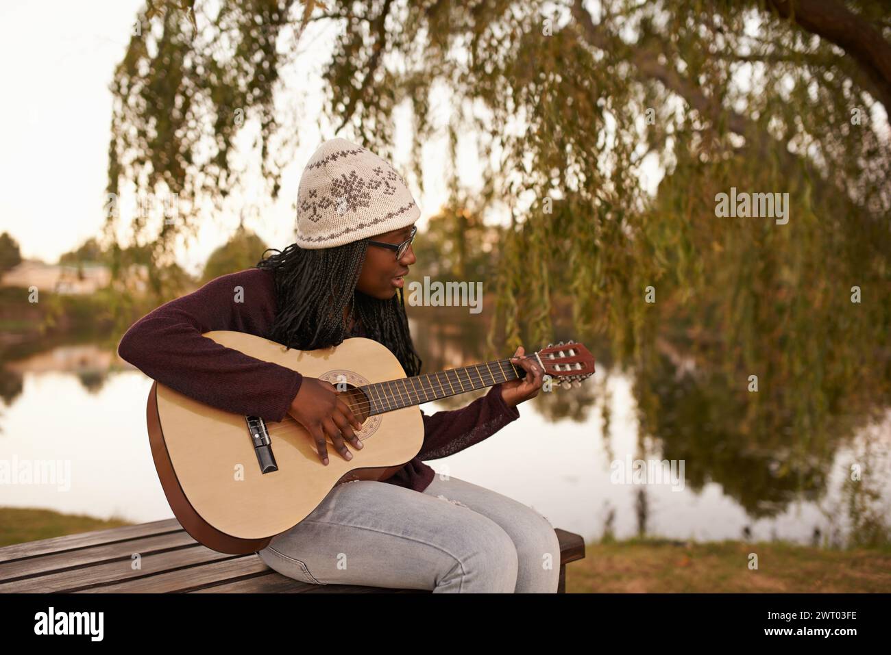 Giovane donna, che suona e chitarra in panchina accanto al lago per la musica, la passione e il talento degli strumenti a corda. Signora africana, musicista e compositore di Song in Foto Stock