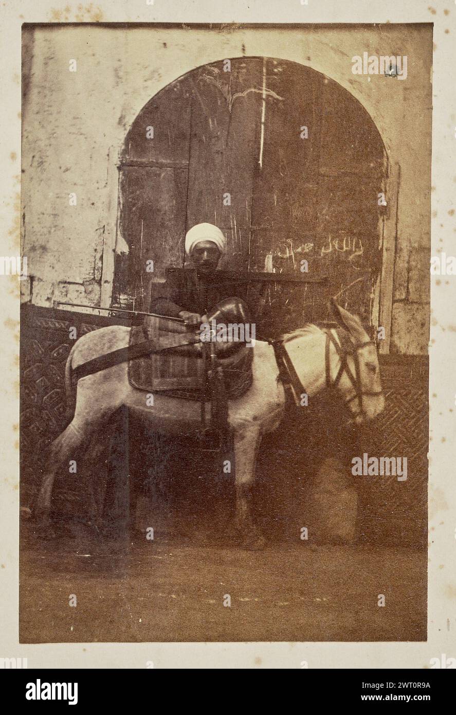 Uomo con un asino. Sconosciuto, fotografo 1861–1903 Un uomo si trova di fronte a una grande porta con un asino. Tenendo un bastone, mette una mano sulla sella dell'asino. La porta reca un'iscrizione in alfabeto arabo. Foto Stock
