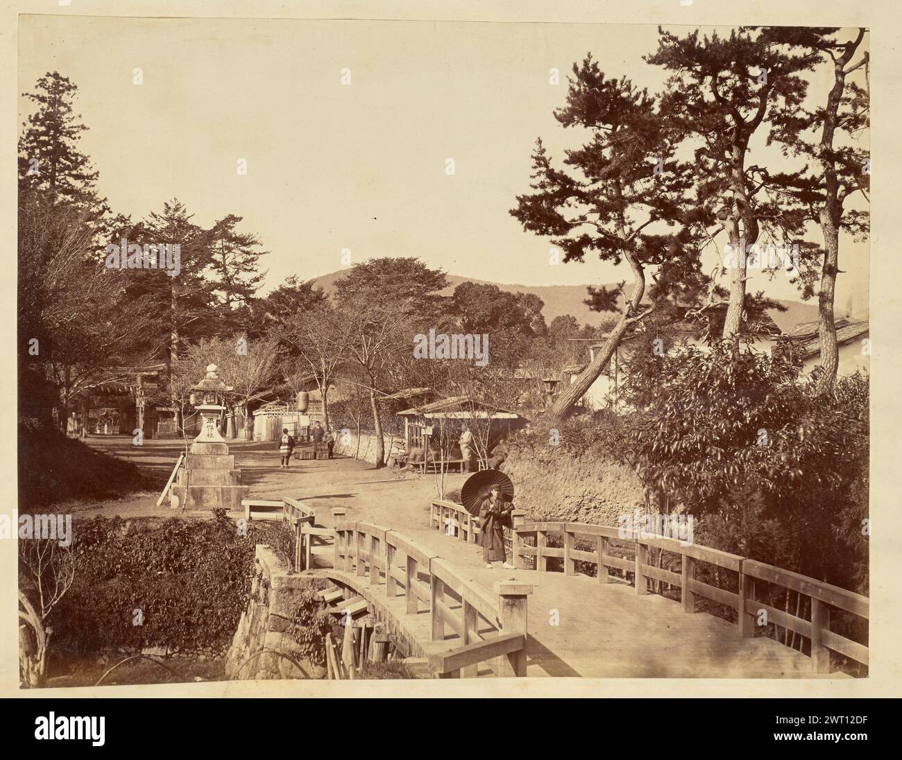 Uomo in piedi su un ponte. Sconosciuto, fotografo anni '1850-'1890 veduta di un uomo in piedi a un'estremità di un ponte che conduce a un piccolo villaggio. L'uomo si appoggia contro un palo di legno nella ringhiera del ponte e tiene un ombrello, o un ombrello di carta ad olio, su una spalla. Un tachidoro, o lanterna a piedistallo in pietra, si trova all'estremità del ponte sul lato sinistro e presenta iscrizioni giapponesi su parte della base. Molte persone si possono vedere muoversi sulla strada sterrata che corre dal ponte attraverso il villaggio. Foto Stock Uomo in piedi su un ponte. Sconosciuto, fotografo anni '1850-'1890 veduta di un uomo in piedi a un'estremità di un ponte che conduce a un piccolo villaggio. L'uomo si appoggia contro un palo di legno nella ringhiera del ponte e tiene un ombrello, o un ombrello di carta ad olio, su una spalla. Un tachidoro, o lanterna a piedistallo in pietra, si trova all'estremità del ponte sul lato sinistro e presenta iscrizioni giapponesi su parte della base. Molte persone si possono vedere muoversi sulla strada sterrata che corre dal ponte attraverso il villaggio. Foto Stock