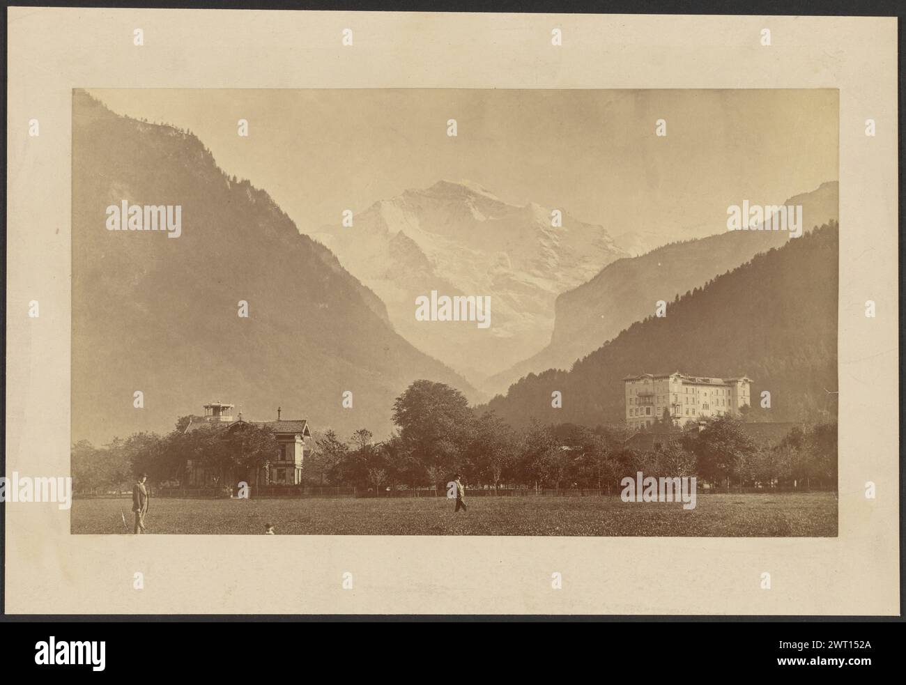 Interlaken Hotel, Svizzera. Sconosciuto, fotografo circa 1870–1890 due uomini in piedi in un campo di fronte a una piccola foresta. C'è una casa con una piccola torre ai margini della foresta e un grande edificio a più piani oltre gli alberi. Gli edifici e gli alberi sono alla base delle montagne. Le montagne più vicine alla foresta sono densamente boscose, mentre una cima in lontananza è ricoperta di neve. (Verso, Mount) centro, matita: "Interlacken Hotel/Jangfraublick [sic]/SWI[illeg]"; Foto Stock