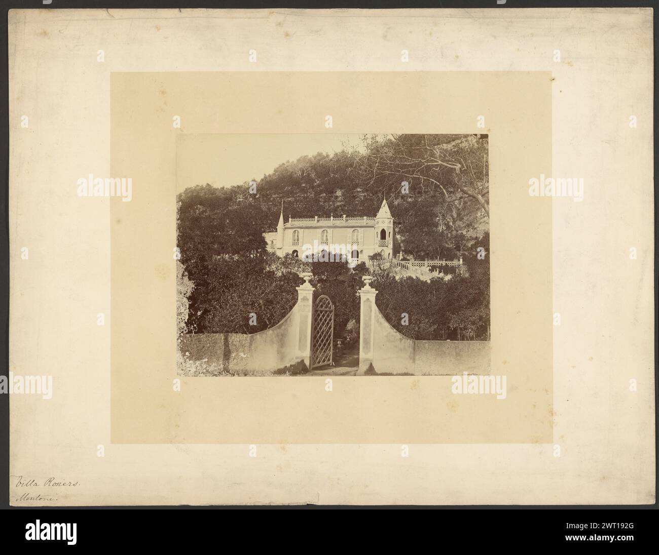 Villa Rosiers, Mentone. Sconosciuto, fotografo 1869 Un palazzo con due torri con tetti a punta, una su entrambi i lati. Il palazzo è visto dal cancello anteriore, adagiato in un basso muro di pietra con un cartello con la scritta "Les Rosiers". Il palazzo è circondato e parzialmente oscurato dagli alberi. (Recto, Mount) in basso a sinistra, inchiostro nero: "Villa Rosiers./Mentone."; (verso, Mount) in basso a sinistra, matita: "Villa R[illeg] Mentone, [illeg] 1869"; Foto Stock