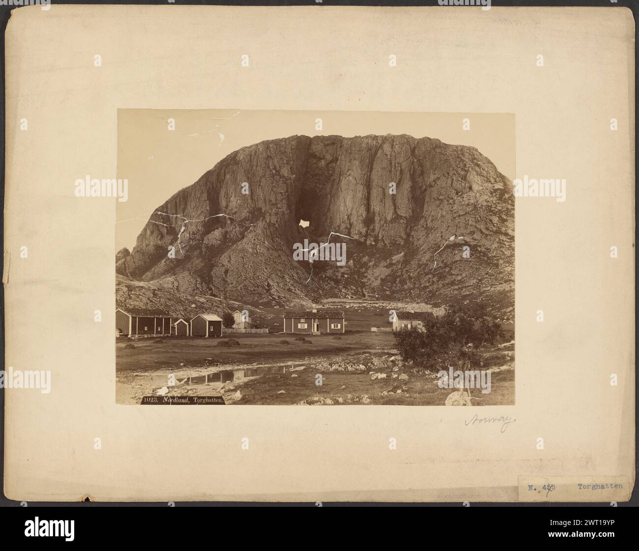 Nordland, Torghatten. Axel Lindahl, fotografo (svedese, 1841 - 1907) circa 1880-1890, Una montagna di granito con un tunnel naturale che forma un buco nel mezzo. Ci sono alcune piccole case nella radura alla base della montagna. La montagna presenta versanti ripidi e un fogliame sparso che cresce lungo la base. (Recto, stampa) in basso a sinistra, stampato in negativo: '1023. Nordland, Torghatten."; (Recto, Mount) in basso a destra, matita: "Norvegia"; in basso a destra, stampa blu su etichetta di carta: "N. 455 ['X' a matita su '455'] Torghatten"; Foto Stock