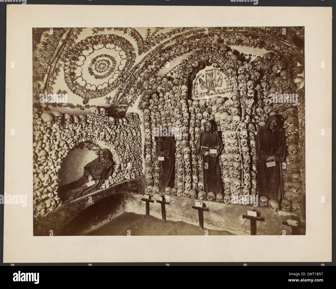 Ossario in una Chiesa dei Cappuccini, Roma. Sconosciuto, fotografo intorno al 1875-1879, Una cripta dove le pareti sono interamente coperte da teschi umani. Ci sono tre scheletri che indossano abiti in posizione eretta e che tengono in mano croci e cartelli. Un quarto scheletro in vesti è posto sulla parete adiacente in una posa reclinata, cullando una croce tra le braccia. (Verso, monte) centro, matita: "Sala Berriale/Chiesa/Capusei [sic]"; Foto Stock