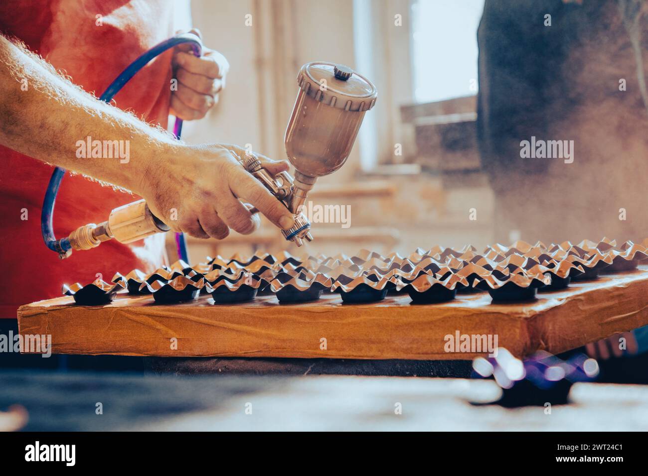 Dipingere oggetti con una pistola a spruzzo in un'officina di verniciatura soleggiata Foto Stock