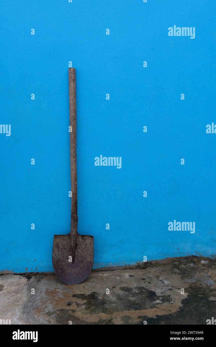 Immagine di natura morta di una pala da giardino a lunga gestione appoggiata su un muro dipinto di blu fuori da una casa rurale del Vietnam. Foto Stock