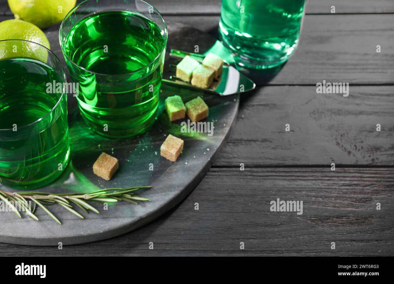 Absinthe in bicchieri, rosmarino, zucchero di canna e lime su un tavolo in legno nero, spazio per il testo. Bevanda alcolica Foto Stock