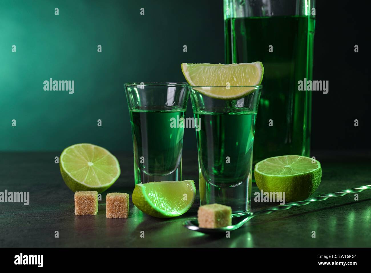 Absinthe in bicchieri da shot, cucchiaio, cubetti di zucchero di canna e lime su un tavolo testurizzato grigio su sfondo verde, spazio per il testo. Bevanda alcolica Foto Stock