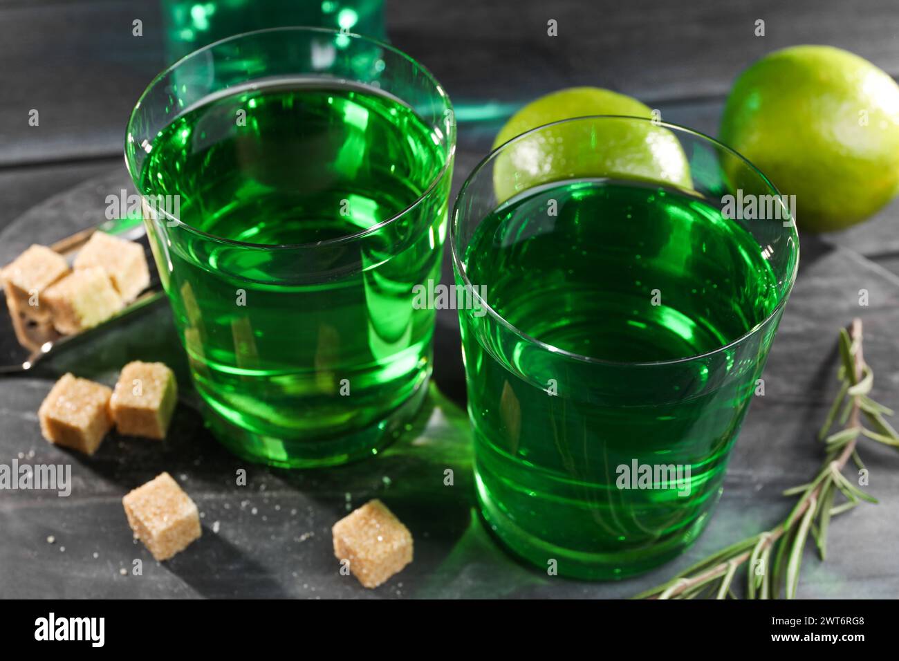 Assenzio in bicchieri, rosmarino, zucchero di canna e lime su un tavolo di legno nero, primo piano. Bevanda alcolica Foto Stock