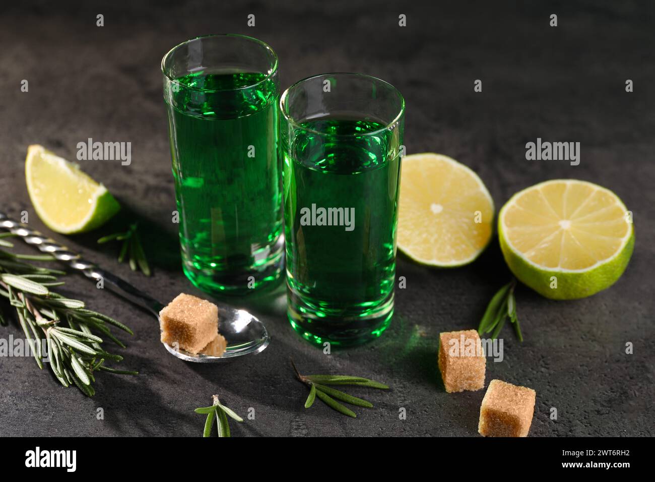 Absinthe in bicchieri da shot, cucchiaio, zucchero di canna, lime e rosmarino su un tavolo testurizzato grigio, primo piano. Bevanda alcolica Foto Stock