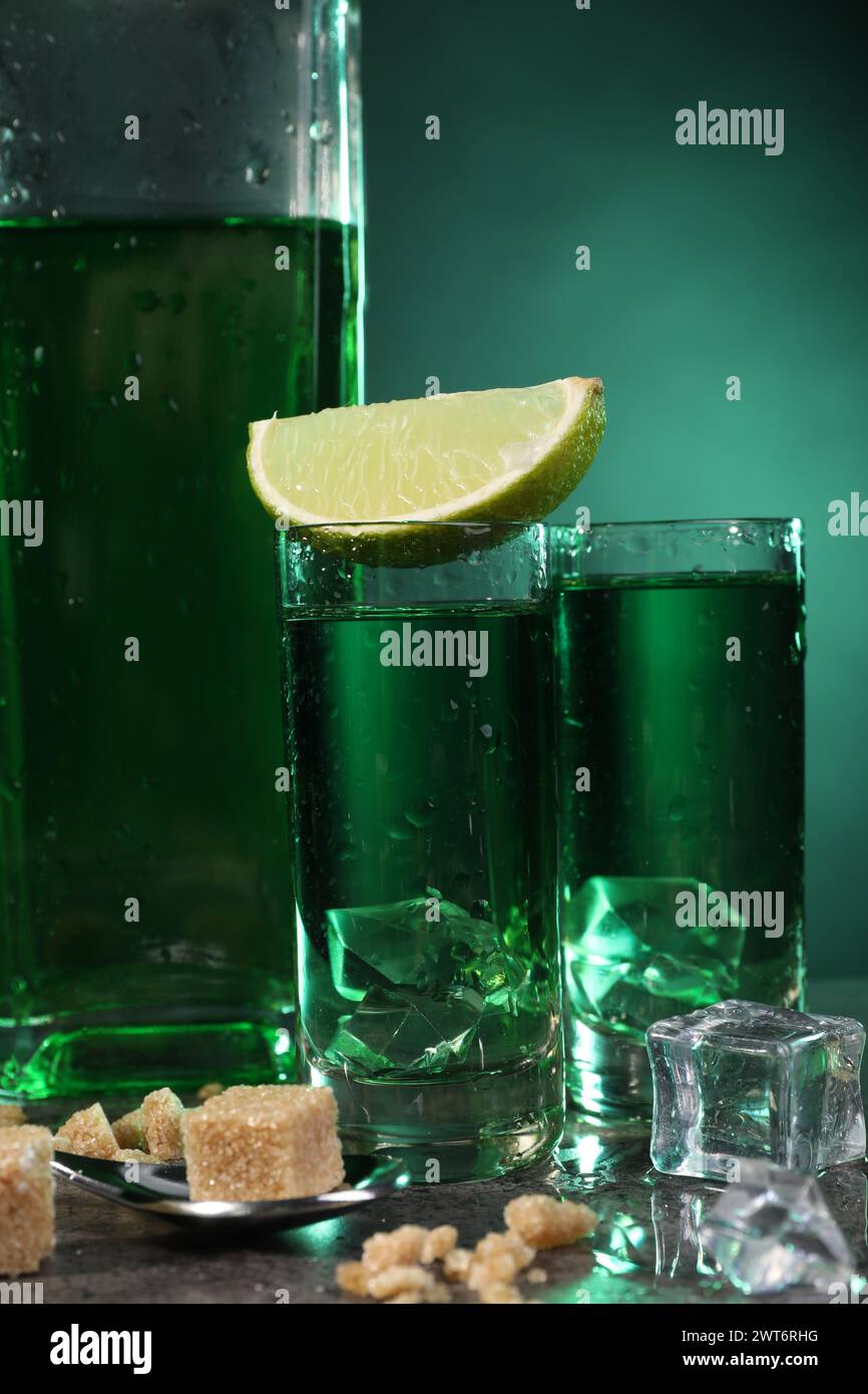 Absinthe in bicchieri da shot con cubetti di ghiaccio, lime, zucchero di canna e cucchiaio sul tavolo grigio, primo piano. Bevanda alcolica Foto Stock