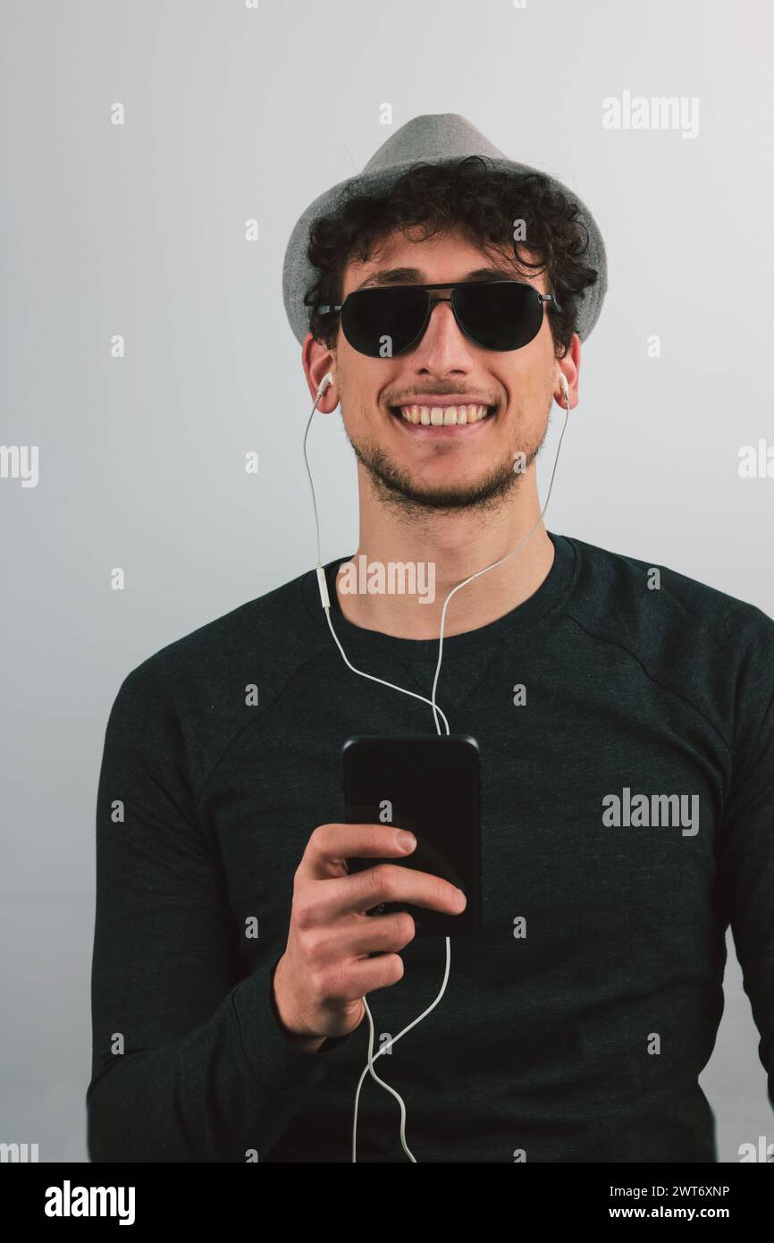 ritratto di un giovane uomo con occhiali da sole con telefono cellulare e in orecchio sorridente su sfondo grigio Foto Stock