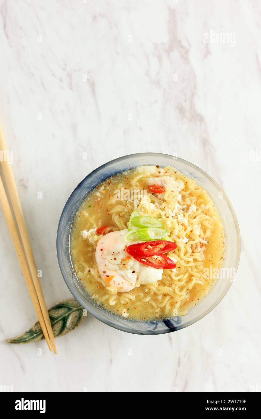 Mie Kuah Telur per Sahur nel Ramadan, zuppa istantanea di spaghetti servita in ciotola di vetro con uovo in camicia e peperoncino. Vista dall'alto su sfondo in marmo bianco. Copia SP Foto Stock