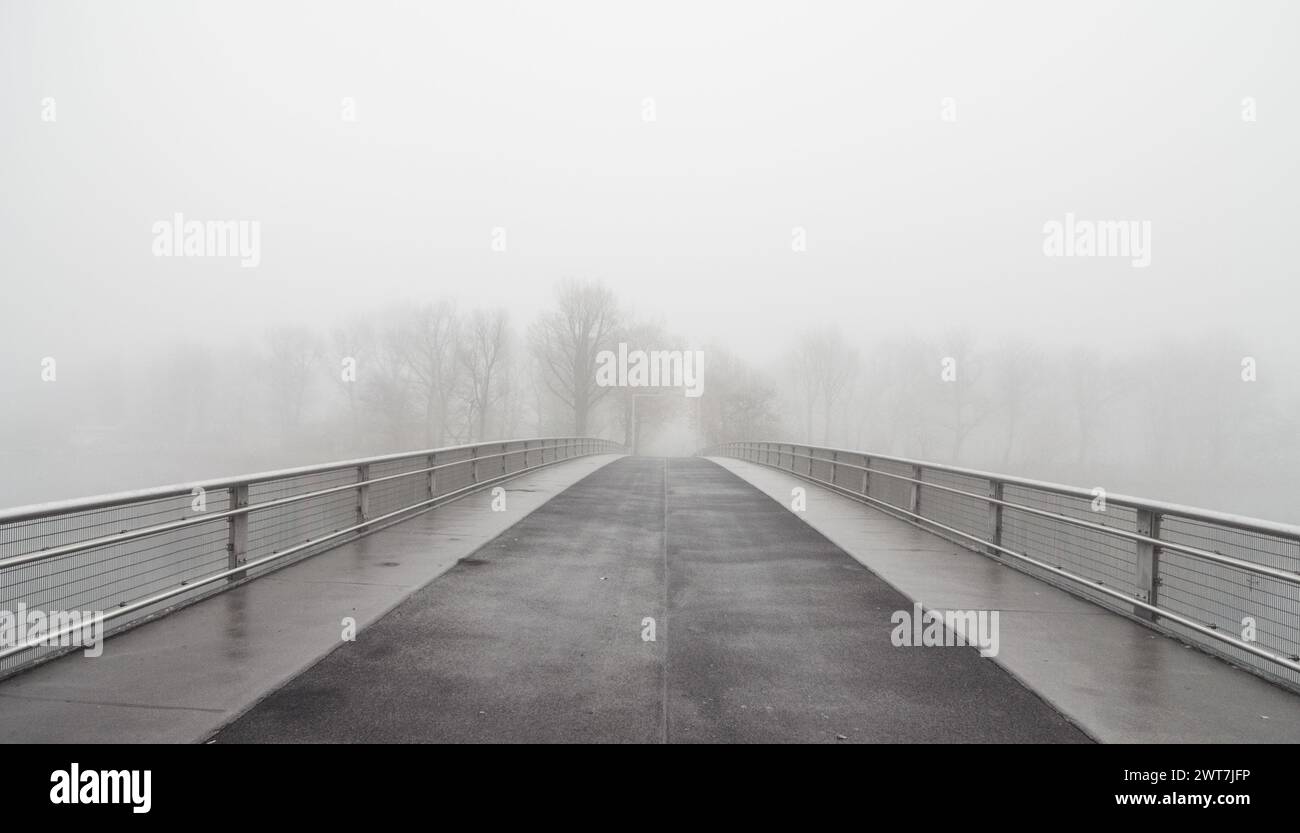 Ponte su un fiume che scompare in una fitta nebbia - bassa visibilità la mattina di novembre. Vista simmetrica del ponte vuoto. Foto Stock