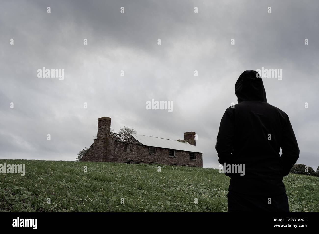 Una misteriosa figura incappucciata che guarda una fattoria abbandonata in rovina su una collina. In una giornata grigia e tempestosa in campagna. Foto Stock