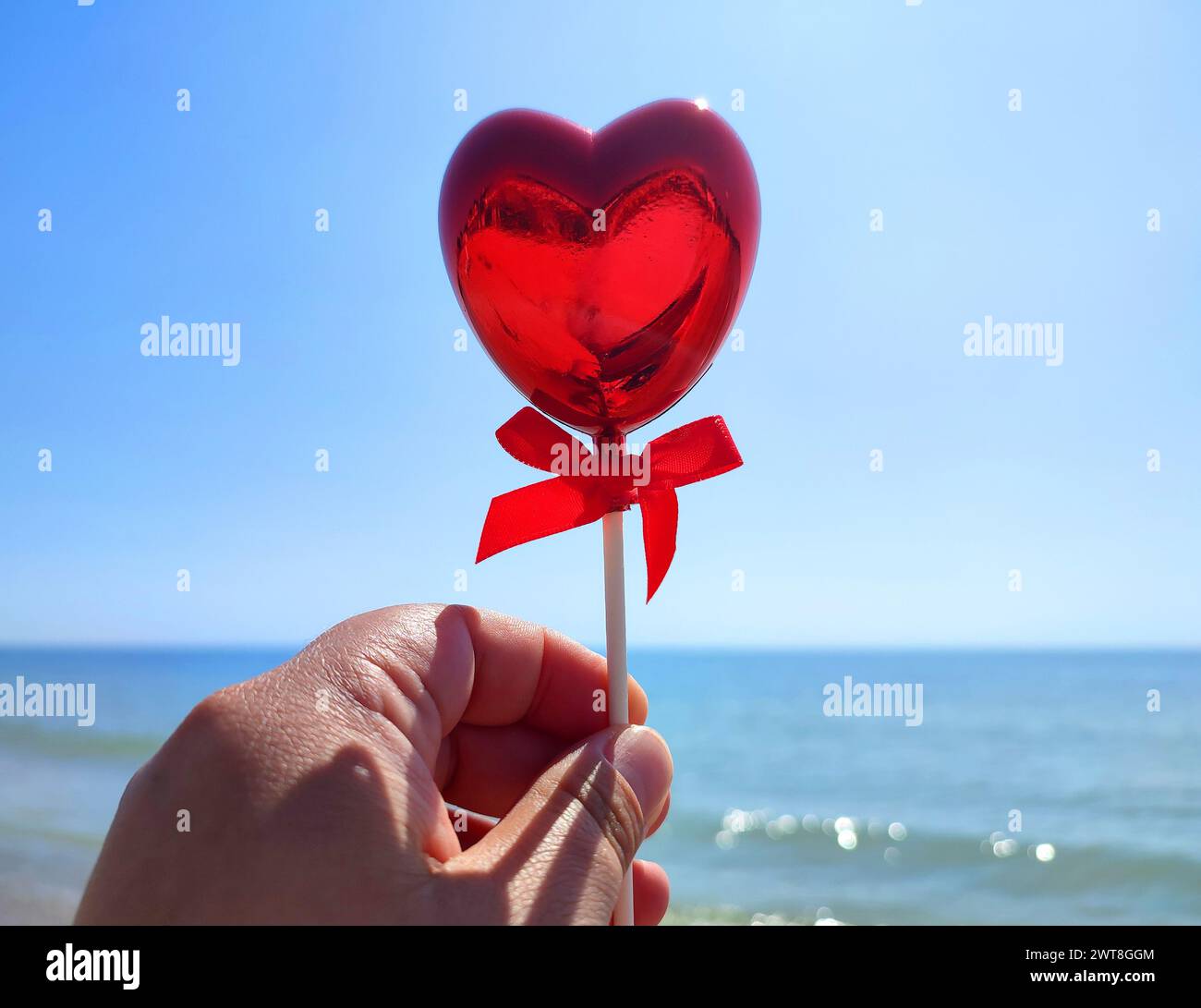 Un bastoncino a forma di cuore rosso volumetrico in mano a una donna sullo sfondo del mare blu, della spiaggia sabbiosa e del cielo blu nelle soleggiate giornate estive. Concetto di amore, San Valentino, innamorarsi. Verticale Foto Stock