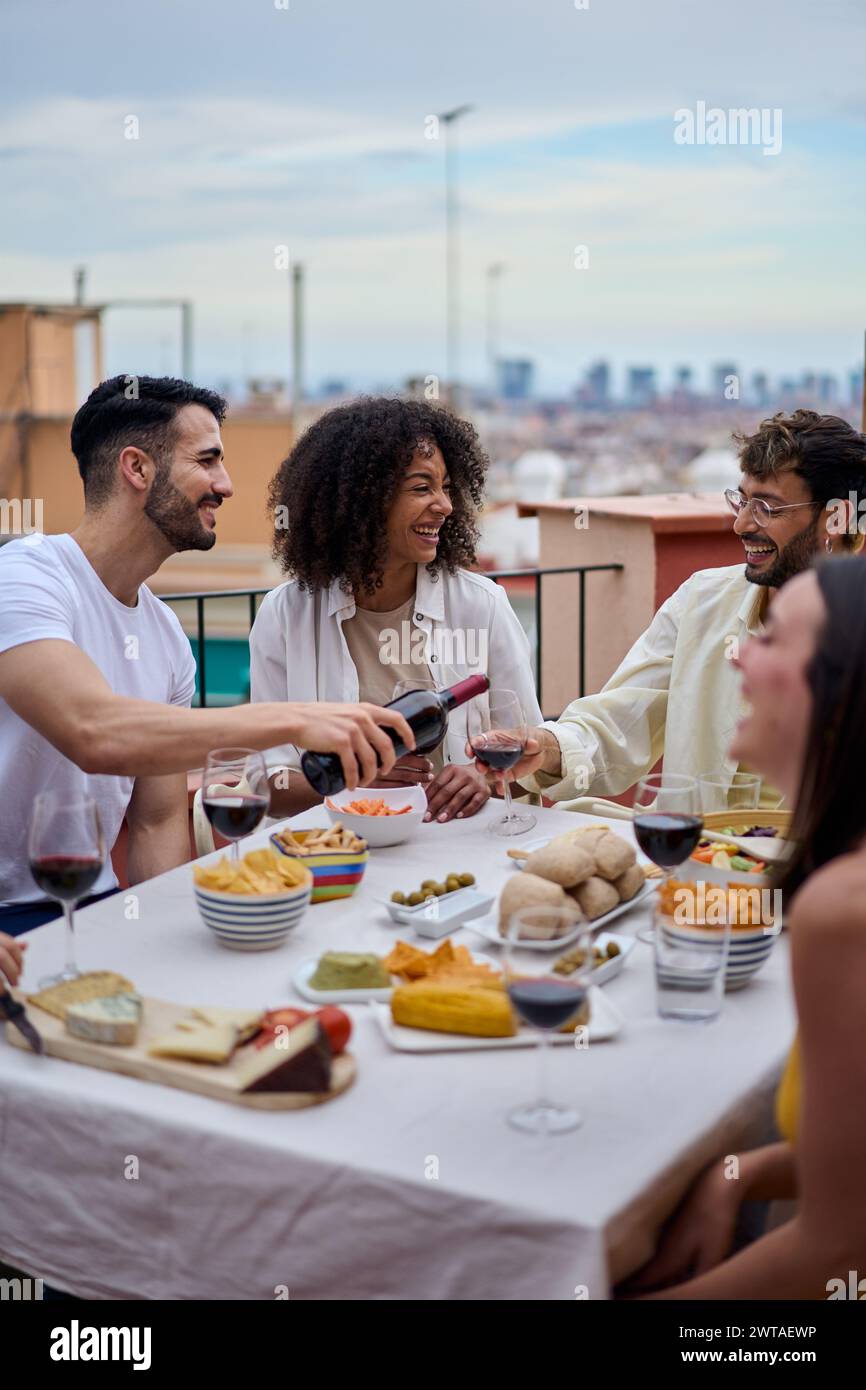 Giovane uomo caucasico sorridente che serve vino rosso agli ospiti al tavolo per festeggiare gli amici felici. Foto Stock
