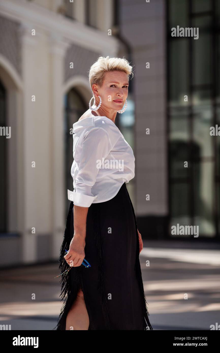 Elegante fashionista di mezza età, vestito con un abbigliamento alla moda gonna nera e una camicetta bianca, passeggia per strada, guardando indietro con eleganza. Bella matur Foto Stock