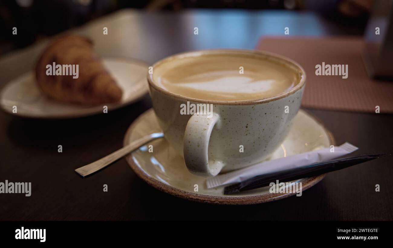 Il delizioso cappuccino italiano caldo si stende in una tazza con le cime di zucchero su un tavolo di legno nel caffè. Sullo sfondo un croissant fresco su un piatto Foto Stock