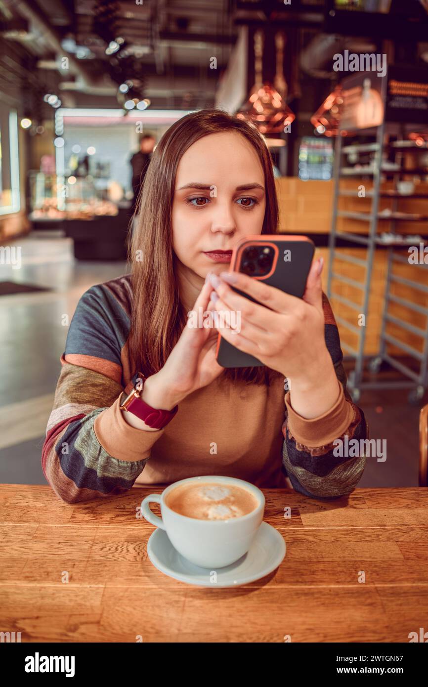 Una donna sfoglia intensamente il suo telefono, una tazza di caffè nelle vicinanze, nell'atmosfera invitante di un caffè locale Foto Stock