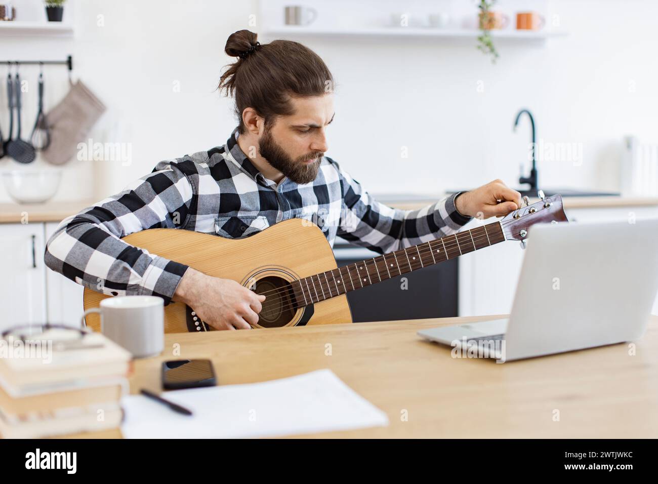 Giovane musicista maschile che sintonizza la chitarra utilizzando un computer portatile sulla scrivania in un appartamento moderno. Foto Stock