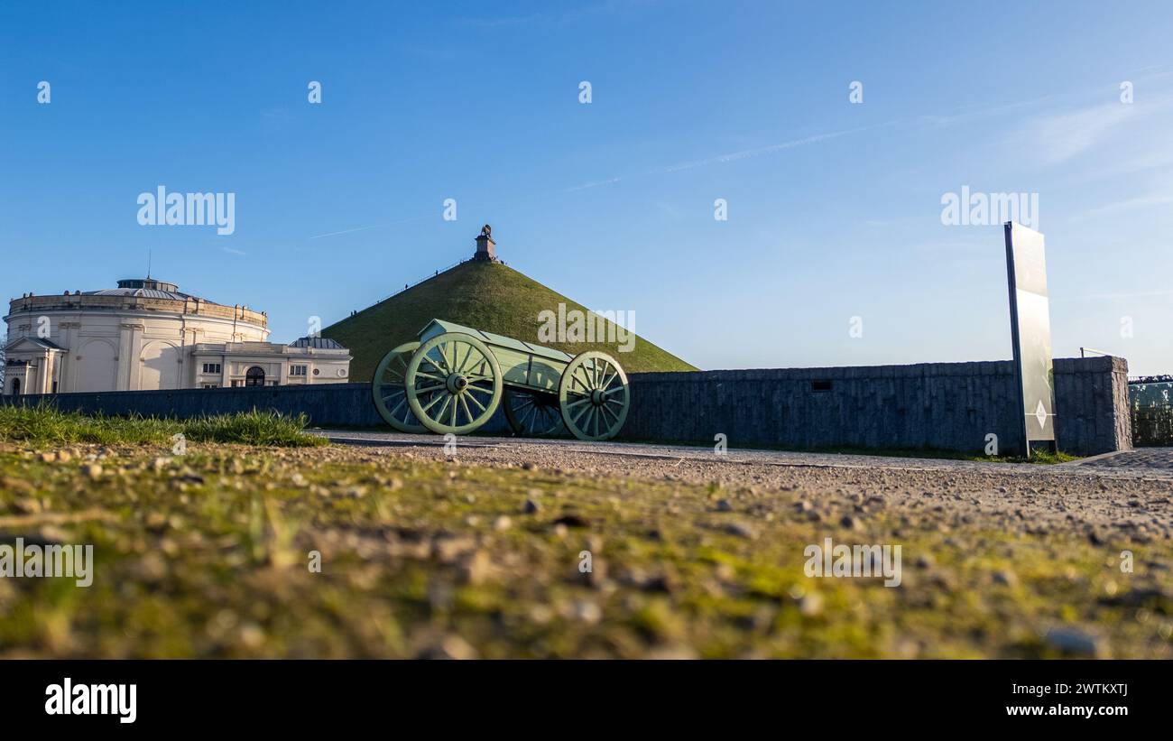 Waterloo, Bruxelles, Belgio, 25 febbraio 2024, questa immagine fornisce una prospettiva unica a livello del suolo di un cannone storico al campo di battaglia di Waterloo, con il tumulo del Leone sullo sfondo. Il cannone è posizionato sulla sinistra, suggerendo il suo ruolo un tempo vitale nella battaglia. Il tumulo del Leone sorge in lontananza, centrato nella cornice con la statua del leone visibile in cima alla sua cima. Il primo piano mostra l'erba ruvida, offrendo un'atmosfera naturale all'ambiente. Il cielo azzurro e limpido sopra dà un tono calmo all'immagine, ricca di simbolismo storico. Vista al piano terra di Waterloo canno Foto Stock