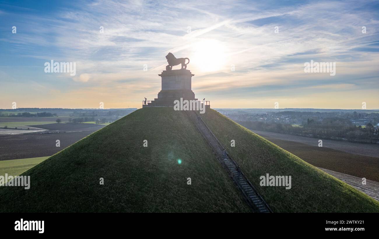 Waterloo, Bruxelles, Belgio, 25 febbraio 2024, si tratta di una tranquilla fotografia all'alba del tumulo dei leoni presso il memoriale del campo di battaglia di Waterloo. Il sole nascente crea un alone luminoso intorno alla statua del leone, che si erge in cima al piedistallo in pietra, creando una silhouette impressionante. I raggi del sole si riversano sul tumulo e sulla campagna circostante, illuminando il sentiero che serpeggia sulla collina. Gli ampi campi sullo sfondo sono immersi nella luce del mattino presto, dando al paesaggio una qualità tranquilla ed eterea. Lion's Mound Memorial silhouette al Sunris. Foto di alta qualità Foto Stock