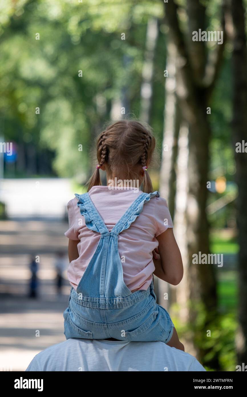 Una giovane ragazza viene portata sulle spalle del padre gode di una giornata di sole in un parco alberato, un'istantanea dell'innocenza e delle semplici gioie del bambino Foto Stock