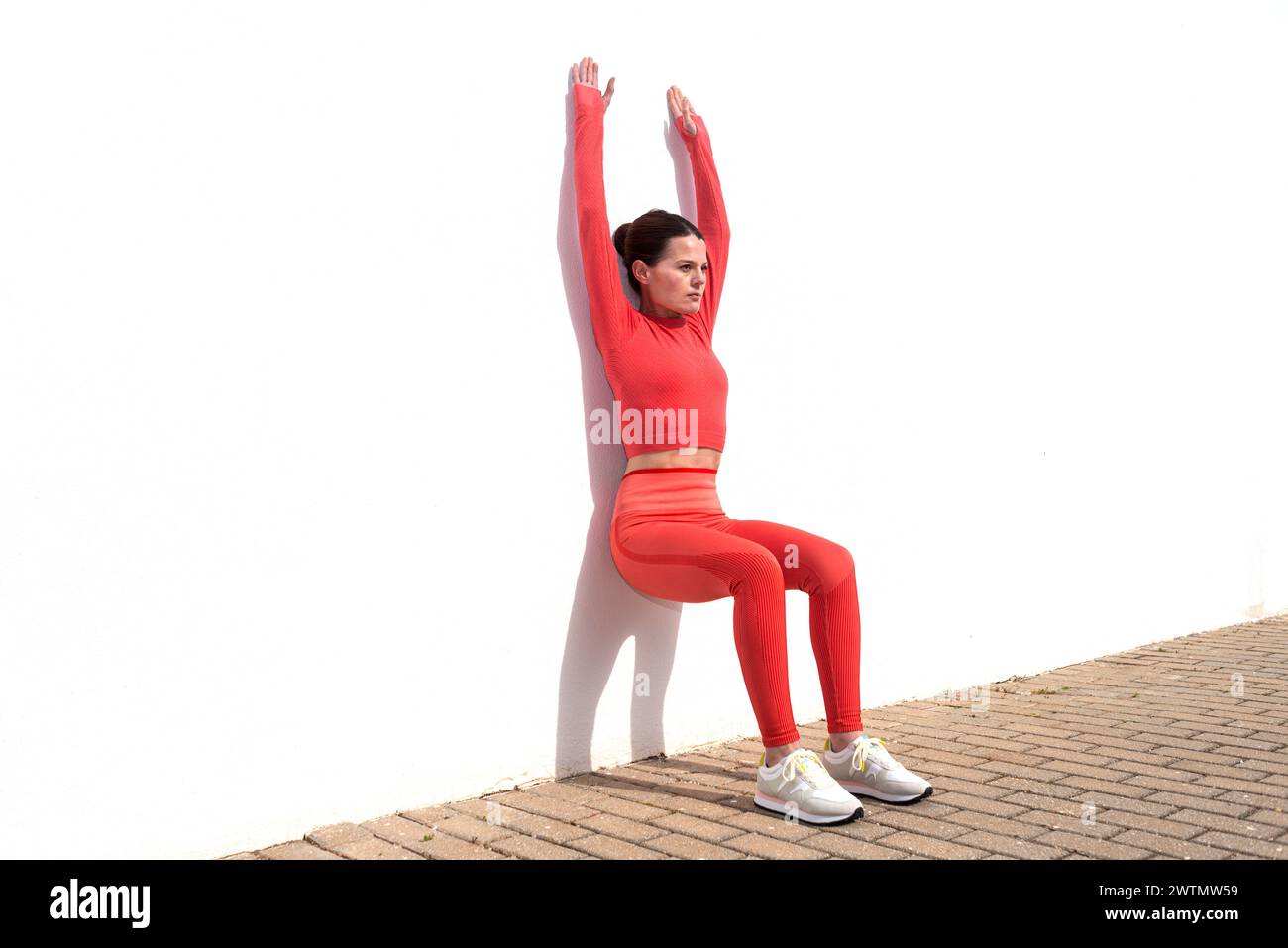 Donna sportiva che fa gli squat contro un muro bianco con le braccia sollevate, fuori al sole. Foto Stock