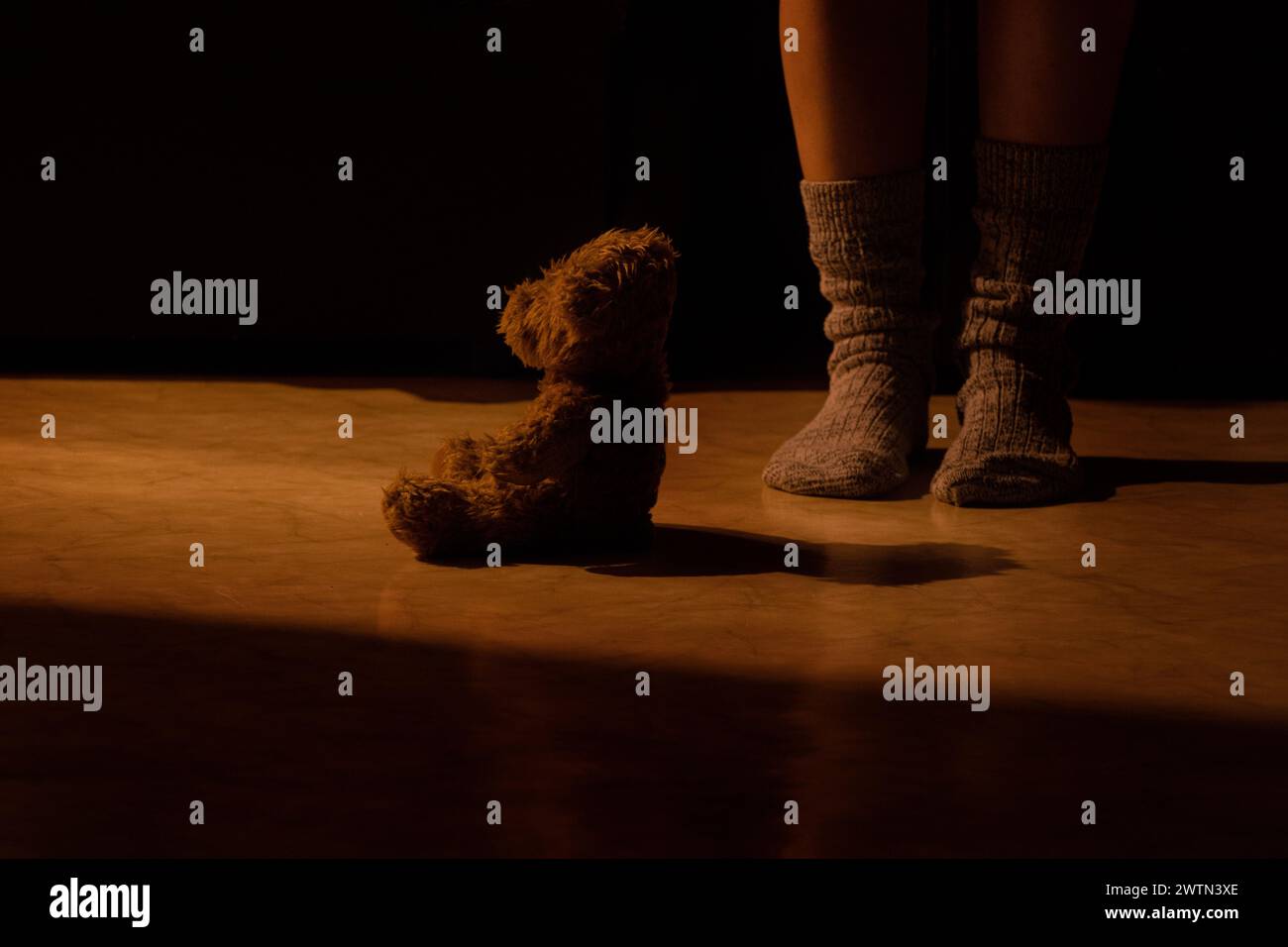 Un orso marrone per bambini siede di notte sul pavimento di una casa in un appartamento e accanto ai piedi delle donne in calze, un giocattolo per bambini Foto Stock