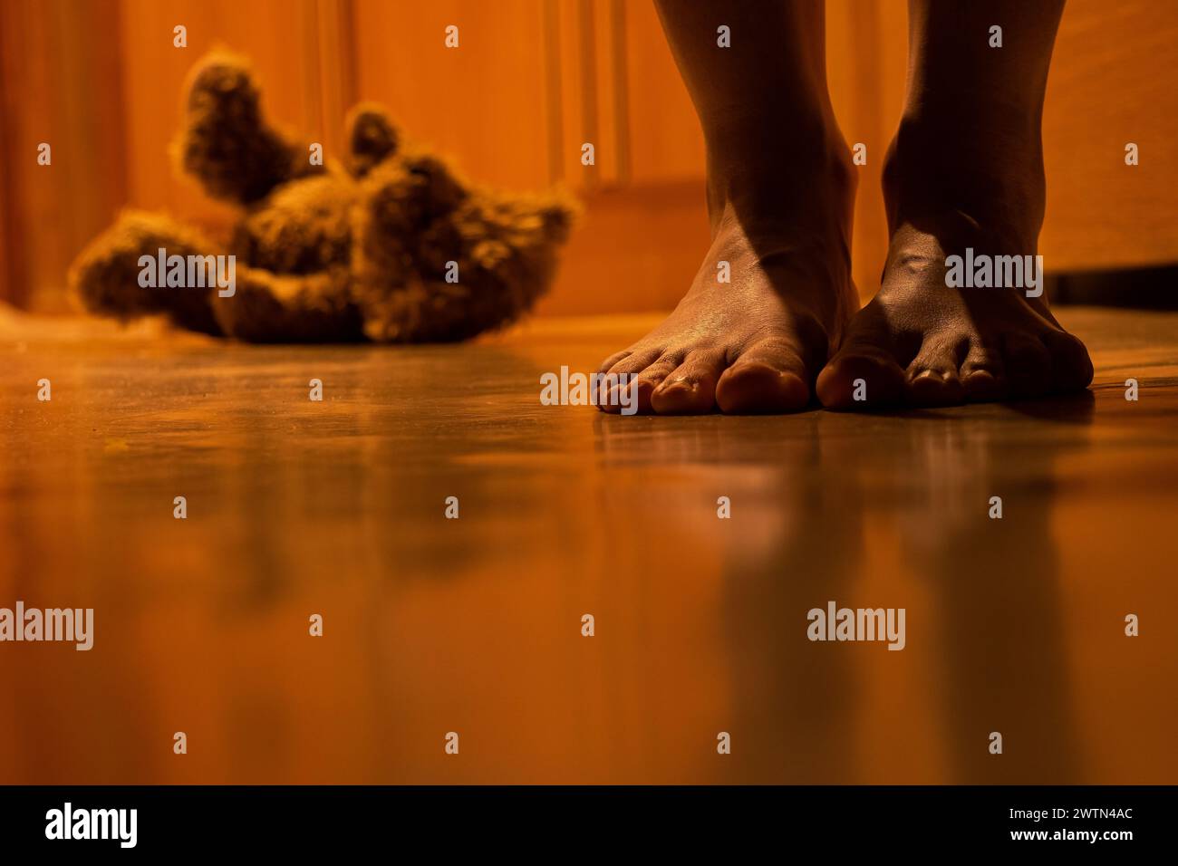 Un orso marrone per bambini si siede di notte sul pavimento di una casa in un appartamento e accanto alle gambe di una donna, un giocattolo per bambini Foto Stock