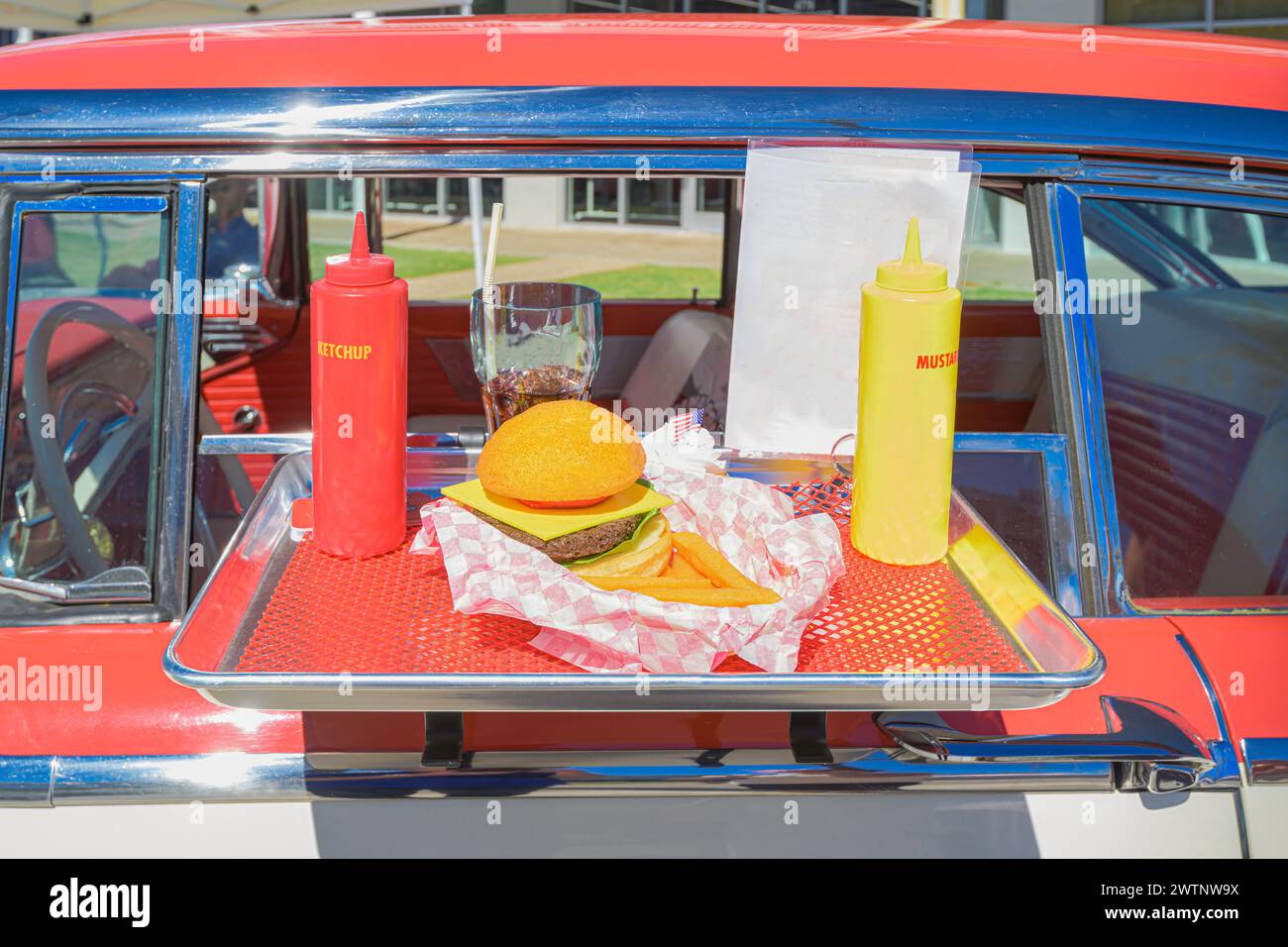 "Classica nostalgia drive-in: Un'auto d'epoca rossa e bianca vanta un vassoio di cibo sul suo finestrino, che mostra una quintessenza americana di cheeseburger Foto Stock