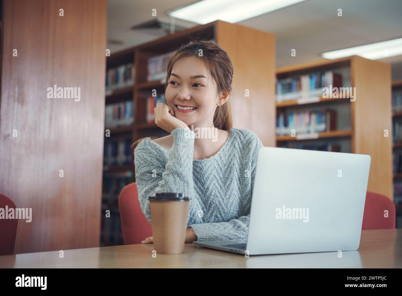 Giovane studentessa felice che studia alla biblioteca del college, seduta alla scrivania, usando un computer portatile. Foto Stock