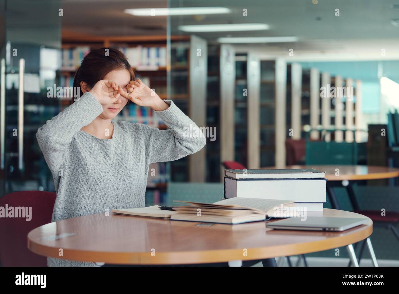 Giovane studentessa asiatica che sfrega gli occhi, si sente stanca dopo aver letto un libro in biblioteca. Foto Stock