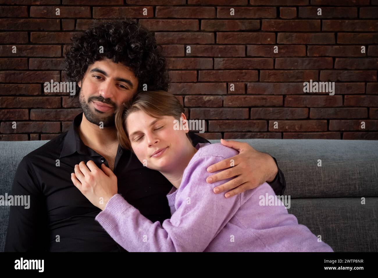 Questa foto mostra un momento tenero tra una coppia che condivide un confortante abbraccio sul divano. L'uomo, con i capelli neri ricci e un'espressione calda, è probabile che sia di origine mediorientale o ispanica. Sta tenendo delicatamente la sua compagna, una donna caucasica con i capelli corti, che sembra serena e rilassata mentre si appoggia a lui, indossando un morbido maglione viola. La parete in mattoni a vista sullo sfondo aggiunge un tocco di fascino rustico alla scena accogliente, aumentando la sensazione di sicurezza e affetto tra i due. Le coppie si abbracciano in un ambiente accogliente. Foto di alta qualità Foto Stock