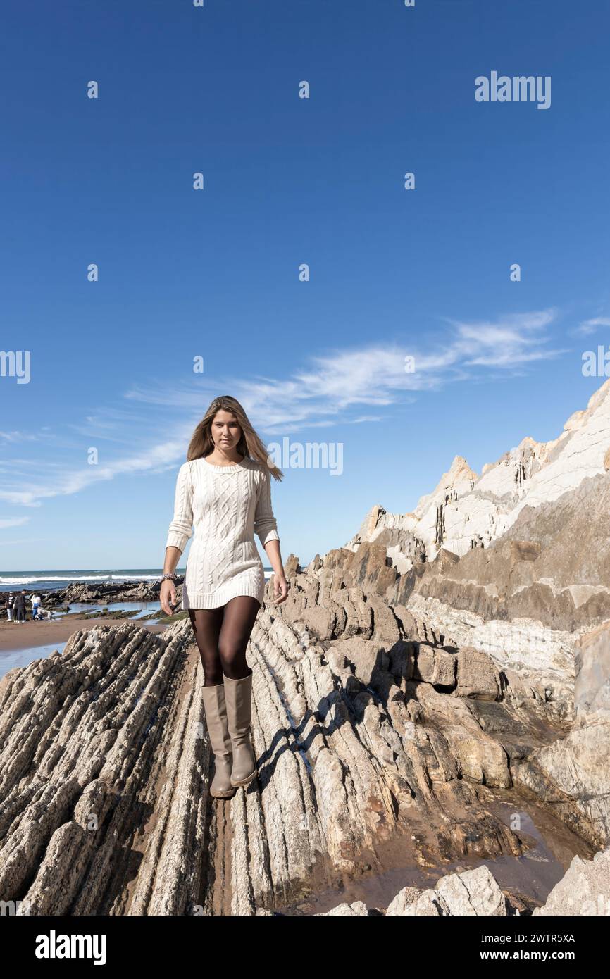 la donna cammina su una spiaggia rocciosa con un cielo azzurro limpido sopra di lei. Indossa un vestito bianco e calze nere Foto Stock
