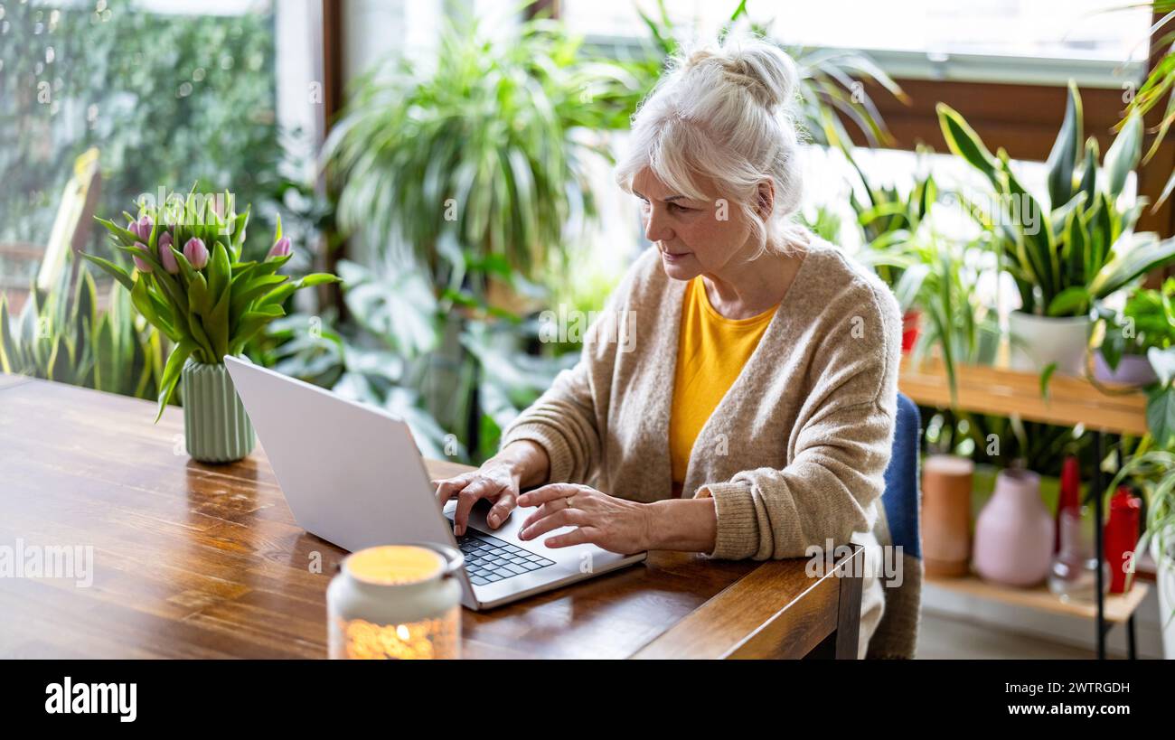 Donna matura che utilizza un notebook seduto al tavolo di casa Foto Stock